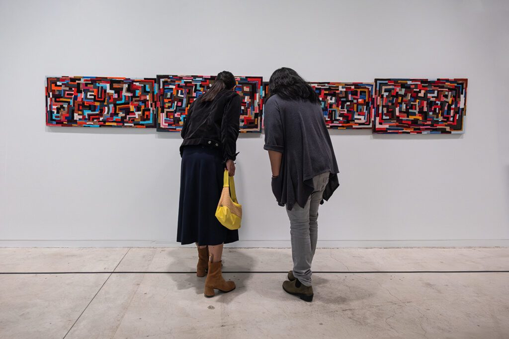 A photograph of two people from behind leaning in to take a closer look at a multicoloured, rectangular work of textile art on the wall in a gallery.
