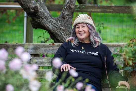 A photograph of artist Dr. T’uy’t’tanat Cease Wyss sitting on a bench in Stanley Park. The background is green, and there is a tree trunk in the back, and flowers in the foreground. The artist is wearing a woven toque and a black sweatshirt.
