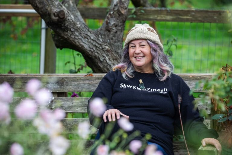 A photograph of artist Dr. T’uy’t’tanat Cease Wyss sitting on a bench in Stanley Park. The background is green, and there is a tree trunk in the back, and flowers in the foreground. The artist is wearing a woven toque and a black sweatshirt.