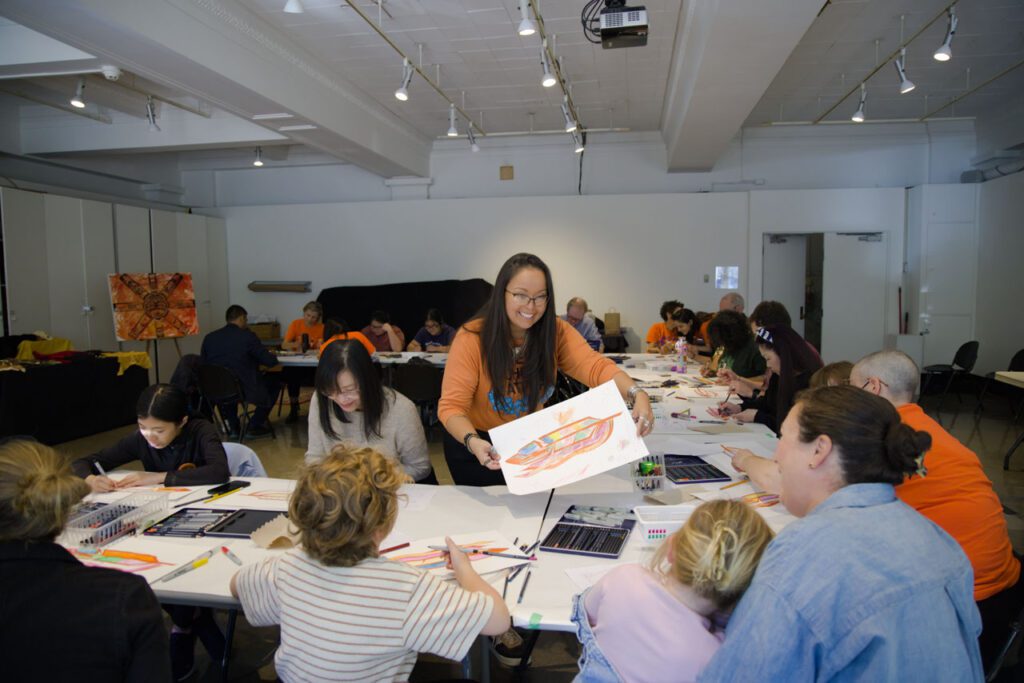 A photograph of an Eagle Feather Design Workshop. There is a room of families sitting at tables arranged in a u shape. An Indigenous woman wearing an orange shirt stands in the centre of the tables and leans over to give an eagle feather drawing to a family.
