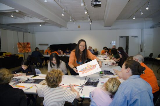 A photograph of an Eagle Feather Design Workshop. There is a room of families sitting at tables arranged in a u shape. An Indigenous woman wearing an orange shirt stands in the centre of the tables and leans over to give an eagle feather drawing to a family.