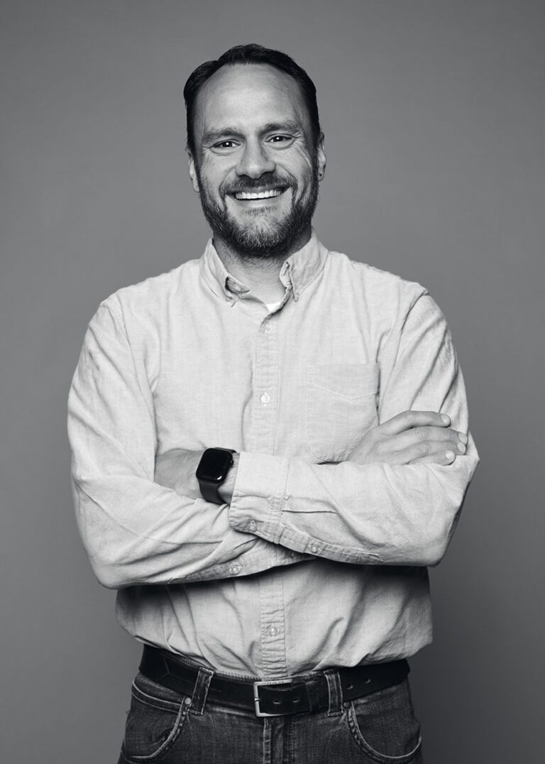 A black and white photograph of a man in a white collared, button up shirt from the waist up. He has a short beard and a large, welcoming smile on his face. His arms are crossed in front of him.