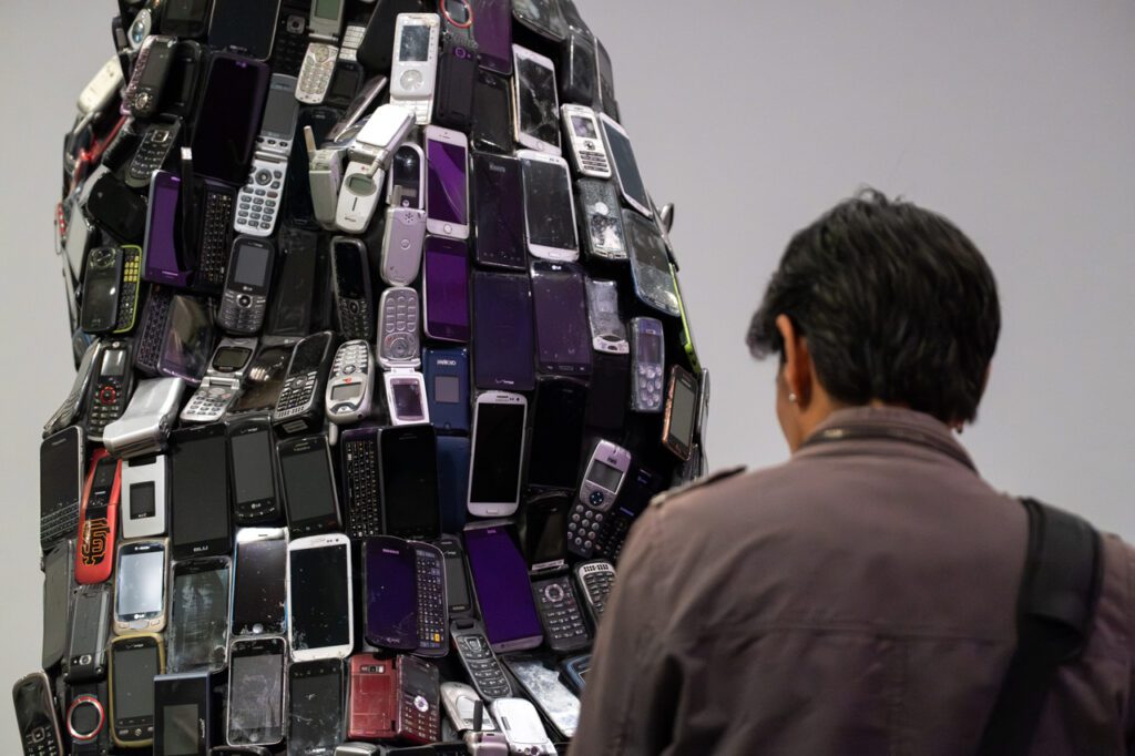 A photograph of a person from behind from the shoulders up. They are looking at a sculpture made up of old cell phone waste.