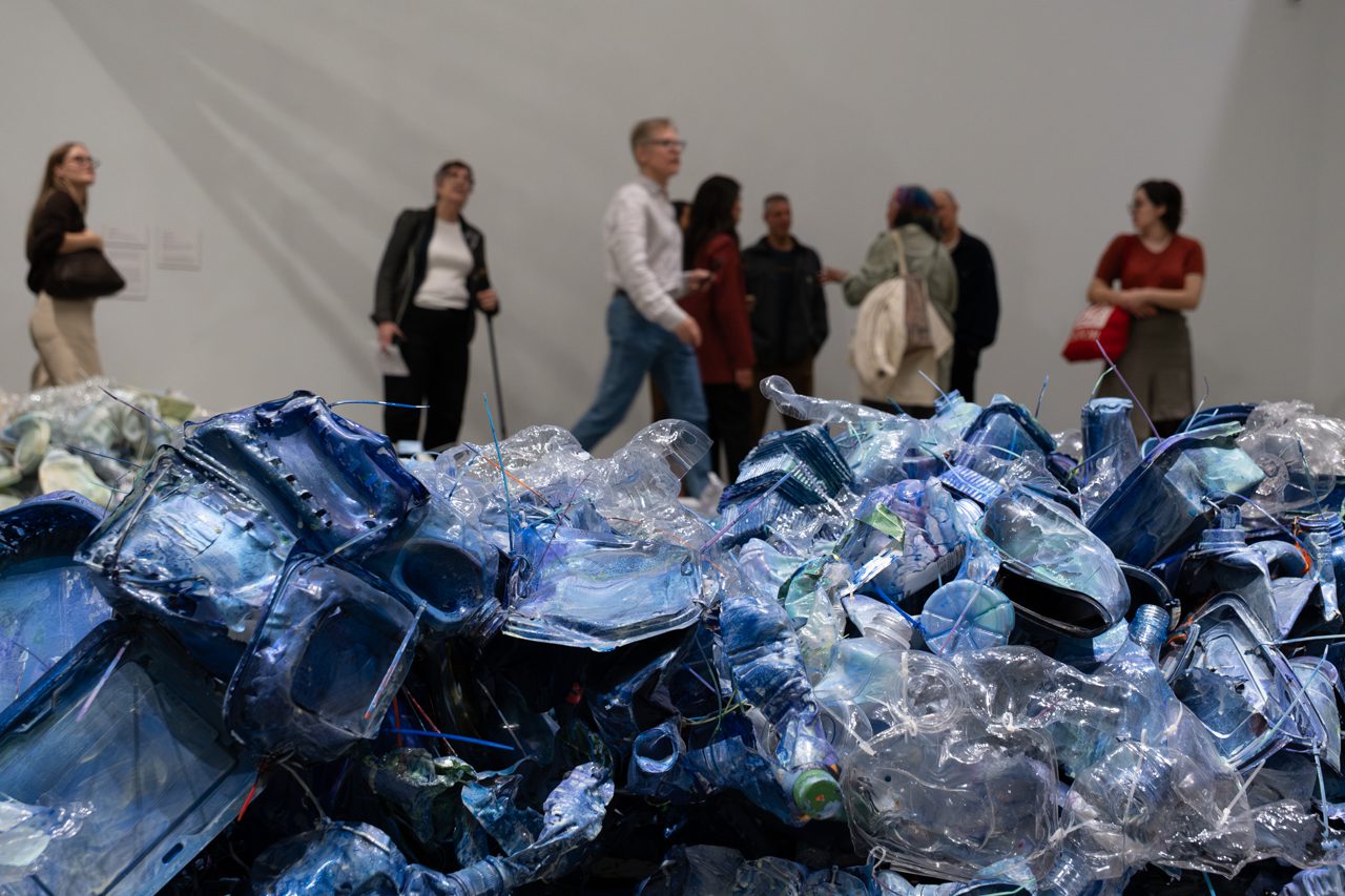 A photograph detailing an artwork by Liz Larner of plastic waste from the ocean, primarily water bottles, painted blue. Behind the wall of plastic in the foreground of the image, there are people mulling about in the gallery space.
