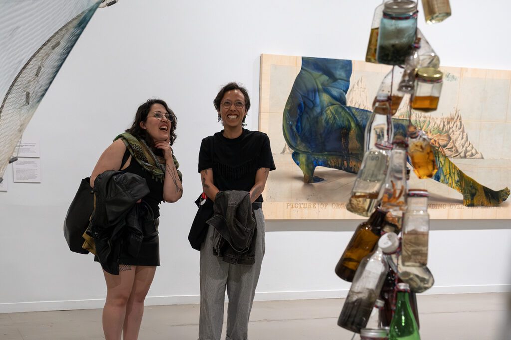 A photograph of two people looking at an artwork made up of bottles with water in them. They are smiling. There is a painting on the wall behind them.