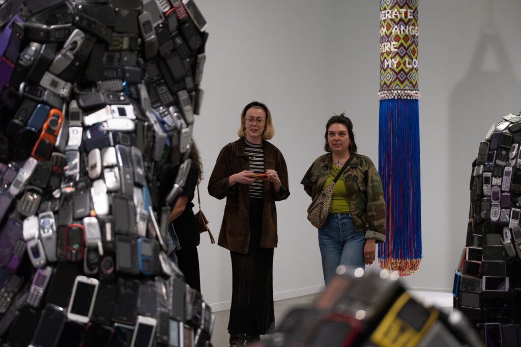 A photograph of two women taking a closer look at a sculpture made up of discarded cell phones.