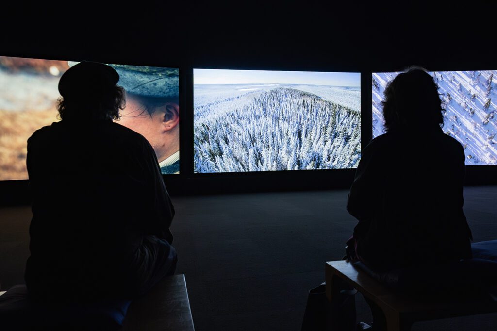 A photograph of two silhouettes of people from behind in a dark room watching a three-channel video projection. In the centre is an image of a mountain covered in snow.