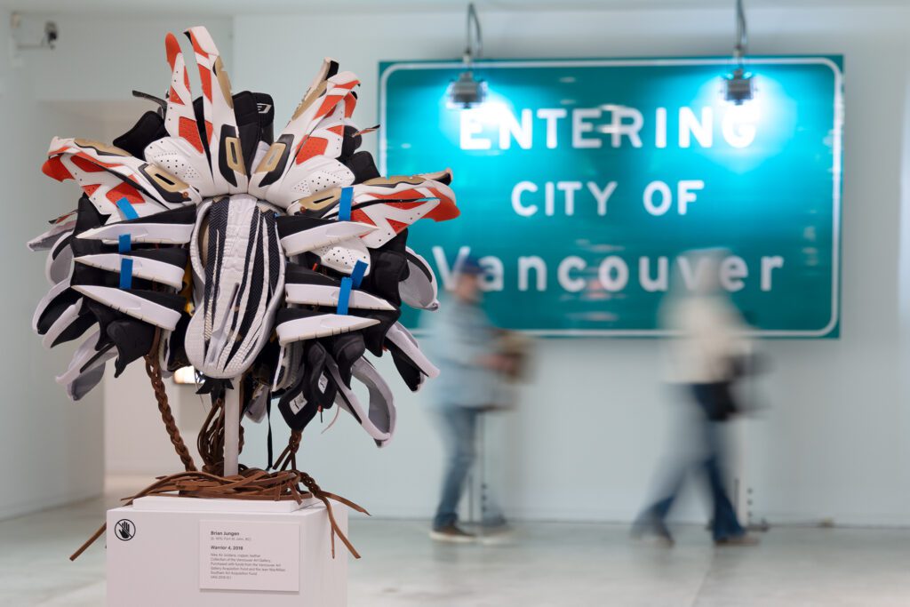 A photograph of a sculpture that looks like an Indigenous mask by artist Brian Jungen. The work is made entirely out of Nike Air Jordan sneakers. In the background, there is a large green sign that reads "Entering the City of Vancouver," with two blurred people walking by.