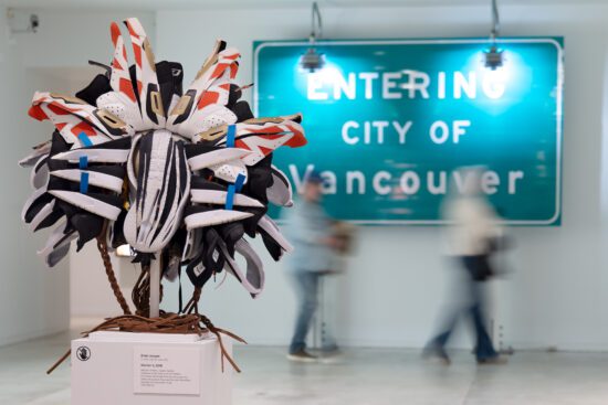 A photograph of a sculpture that looks like an Indigenous mask by artist Brian Jungen. The work is made entirely out of Nike Air Jordan sneakers. In the background, there is a large green sign that reads "Entering the City of Vancouver," with two blurred people walking by.