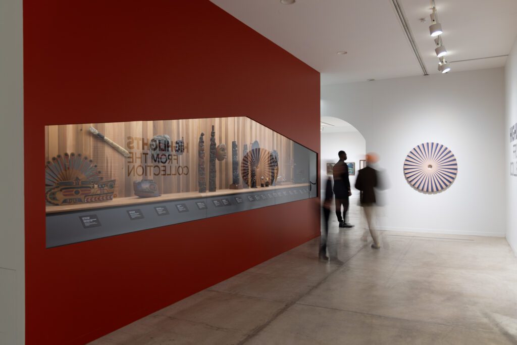 A photograph of the opening room in the exhibition Highlights from the Collection. On the right is an enclosure on a red wall with glass, and in it are Indigenous artworks. On the far wall on the left is a circular pink sculpture.