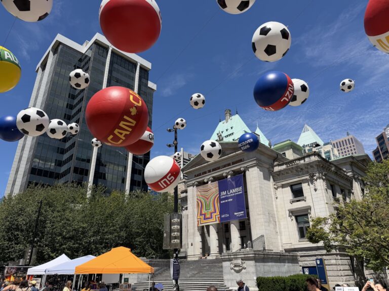 A photograph of the exterior of the Vancouver Art Gallery from Robson Street Plaza. There are black and white and colourful soccer balls seemingly floating in the air as part of an installation. The neo-classical Gallery building is visible behind. There is a banner advertising an exhibition of Jim Lambie's work on the building facade.