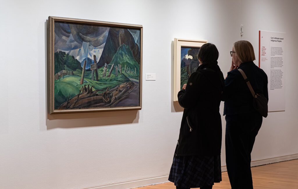 Two visitors stand side by side in a gallery, closely viewing framed paintings on a white wall. The central artwork is by Emily Carr, depicting a stylized landscape with green hills, fences and dramatic, swirling clouds, surrounded by other paintings and wall text.