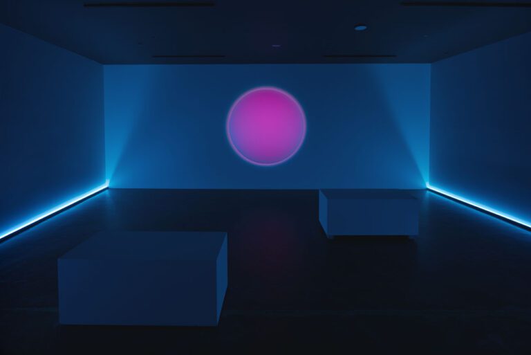 A photograph of an artwork by Annie Briard. The image shows a darkened room that has a blue hue. There are dim lights along the walls, and the outline of benches are visible. In the centre is a large, circular pink orb that appears to glow.
