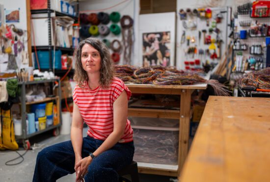 A photograph of artist Emily Hermant sitting in her studio. She sits on a stool off to the left of the image. She is wearing a striped pink shirt and dark wash denim jeans. She looks out at the camera. She has brown, curly hair. Behind her is her studio full of wires and technological waste used as part of her practice.