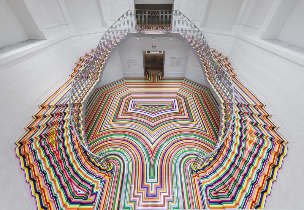 An installation photograph of Jim Lambie's artworks Zobop (Colour-Chrome) and Zobop (Colour-Chrome) Stairs covering the stairs and floor of the Vancouver Art Gallery's neo-classical Rotunda. The floor and stairs are covered in brightly coloured strips of vinyl in purple, pink, green, red, orange and yellow.