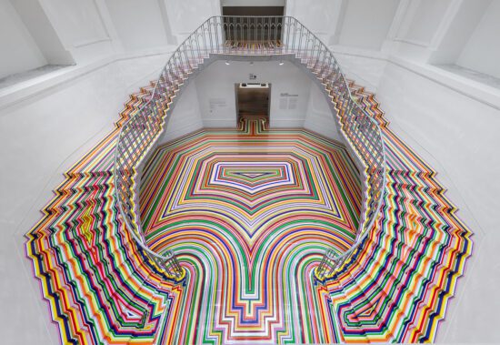 An installation photograph of Jim Lambie's artworks Zobop (Colour-Chrome) and Zobop (Colour-Chrome) Stairs covering the stairs and floor of the Vancouver Art Gallery's neo-classical Rotunda. The floor and stairs are covered in brightly coloured strips of vinyl in purple, pink, green, red, orange and yellow.