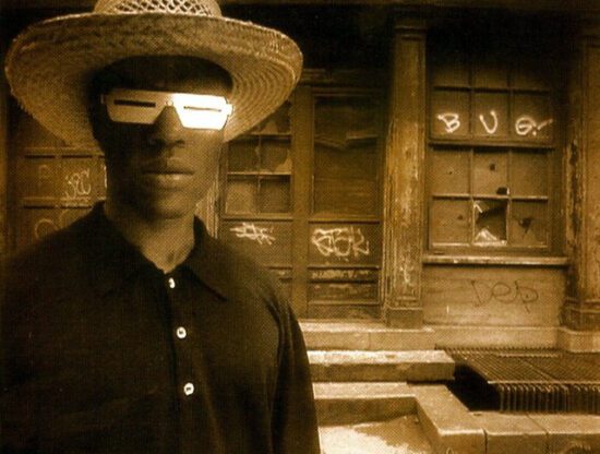 A film still from The Last Angel of History. The image is pixelated and in a sepia tone. A Black man wearing a hat and metalic sunglasses with small slits in them appears on the left. Behind him is a building that has graffiti on it.