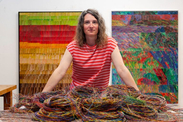 A photograph of artist Emily Hermant in her studio. In the image, a woman with brown, curly hair in standing at the centre with her arms on a table covered in colourful wires. In the background, there are two artworks that look like abstract paintings from a distance.