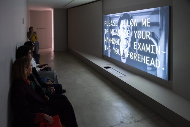 A photograph of a darkened room with a video projected on a screen. A group of people sit against the wall.
