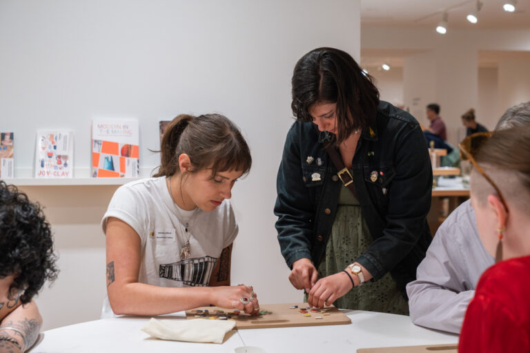 A photograph of two women in an exhibition space. One is sitting at a table, and the other is standing, looking down at her hands. They are both working on an activity.