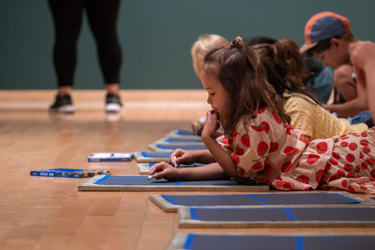A photograph taken from the side of a group of children drawing in the Gallery. In the foreground, a girl in a dress covered in a tomato print looks thoughtfully at her paper.