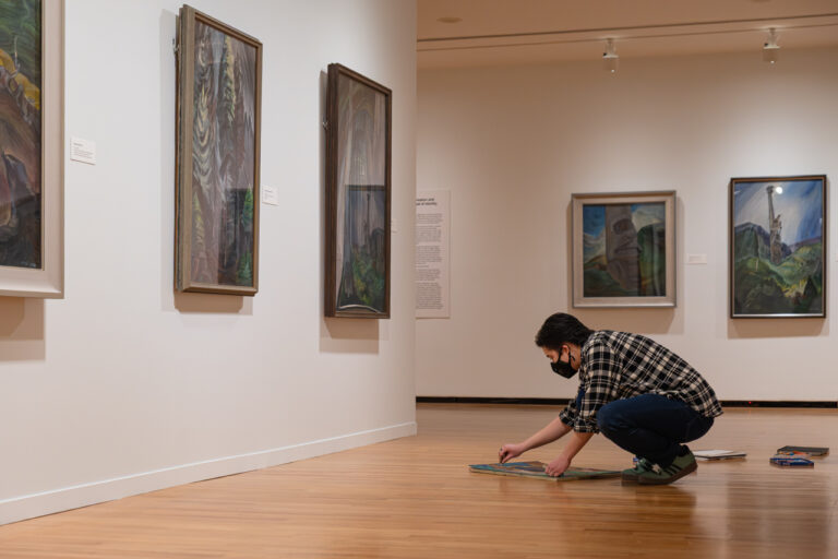 A photograph of a teenager sitting on the floor in the Emily Carr exhibition space surrounded by paintings. They are drawing.