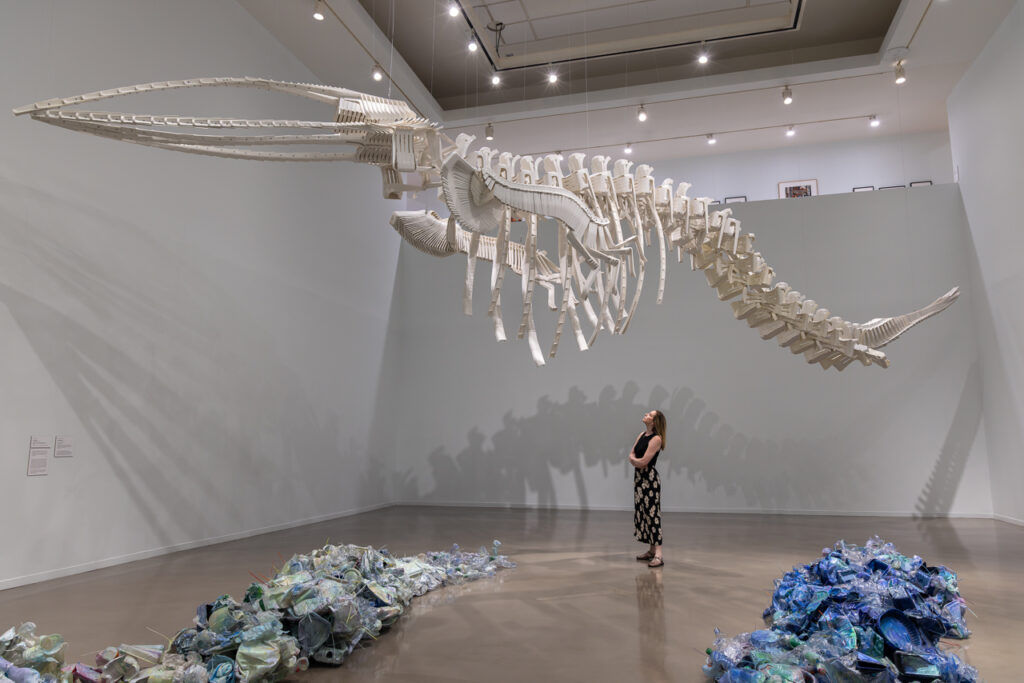 A photograph of a blond-haired woman standing beneath a large whale skeleton made of white plastic lawn chairs. On the floor are discarded plastic bottles and items painted blue.