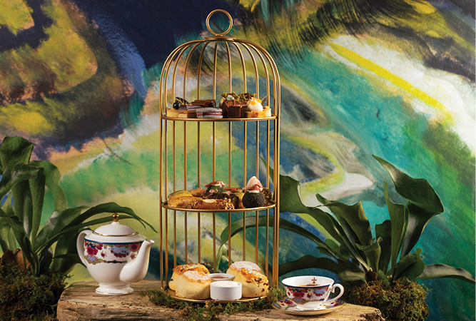 Afternoon tea display featuring a tiered tray of pastries and sandwiches, a floral teapot and teacup, set among greenery against a colorful painted backdrop inspired by Emily Carr