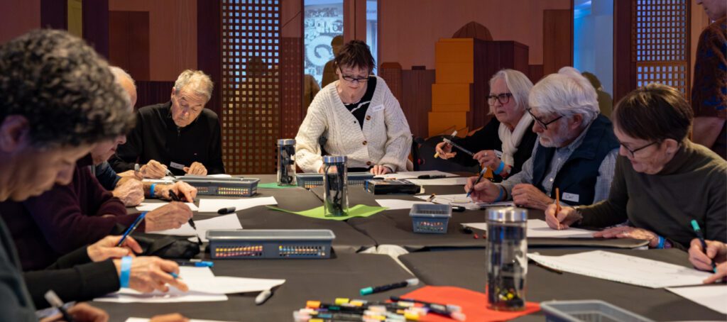 A photograph of a group of seniors, men and women, seated at a table during a workshop. They are drawing.