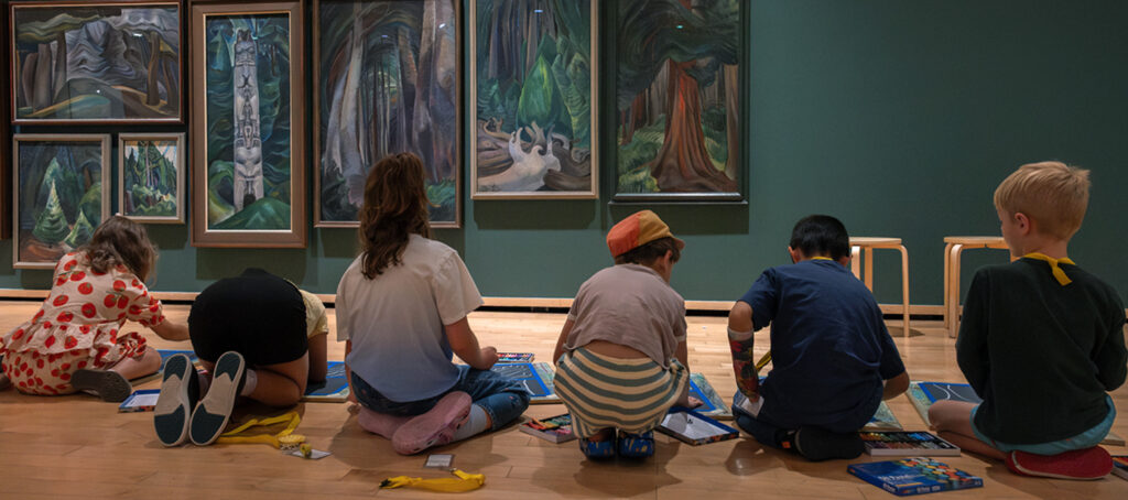 A group of children sit in the Gallery in front of a wall of Emily Carr paintings of trees and draw.