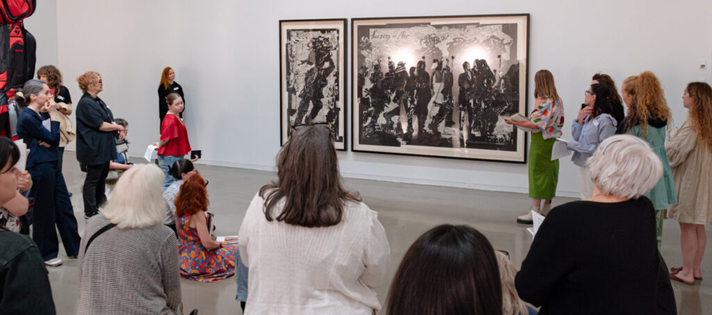 A photograph of a group of approximately 10 women from the back standing in front of an artwork and looking intently.