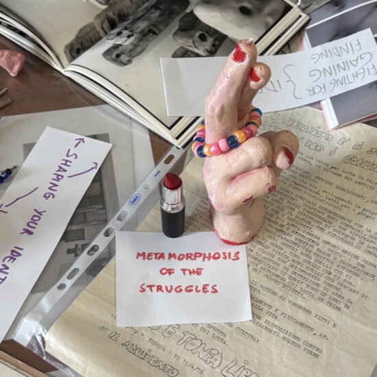 A photograph of a display hand with red nail polish crossing it's finders. Papers and archival materials surround it, along with a red lipstick.