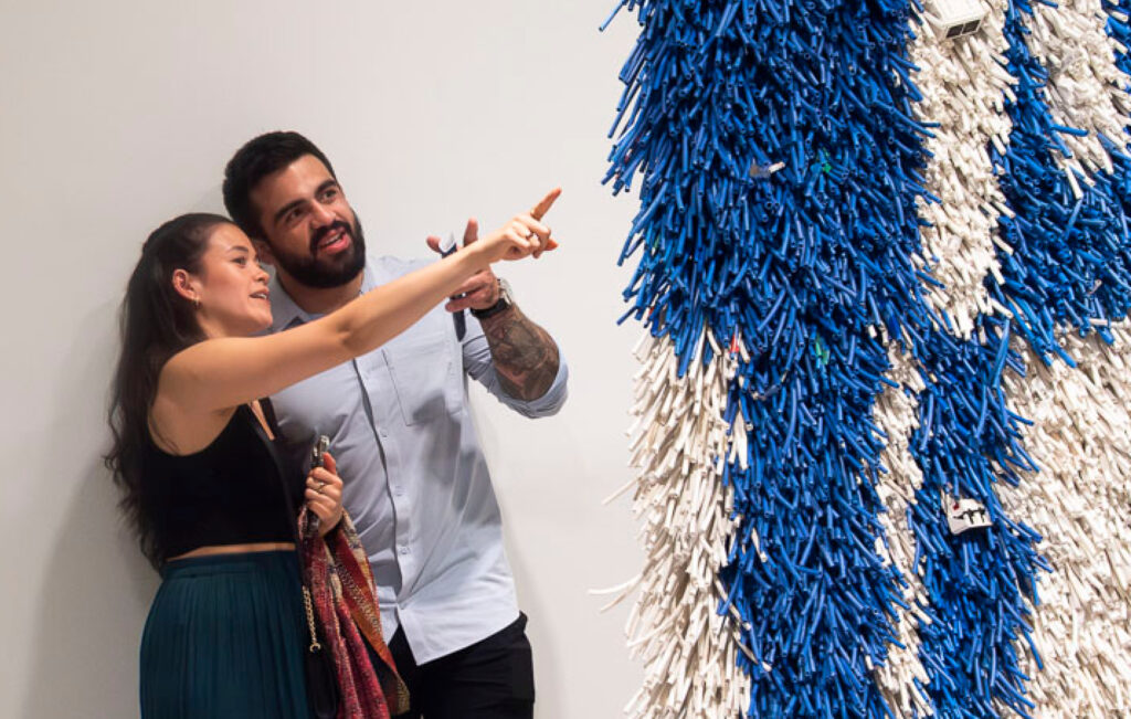 A photograph of a woman and a man looking at a blue and white artwork that looks like a hanging tapestry. The woman points at the work as she discusses it with her friend.